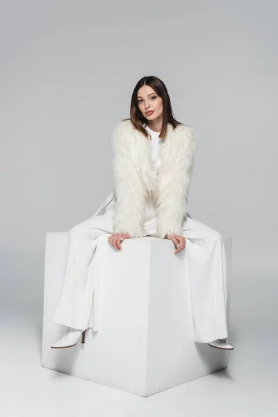 Volle Länge der stilvollen Frau in Kunstpelzjacke und insgesamt weißes Outfit sitzt auf Würfel auf grau — Stockfoto