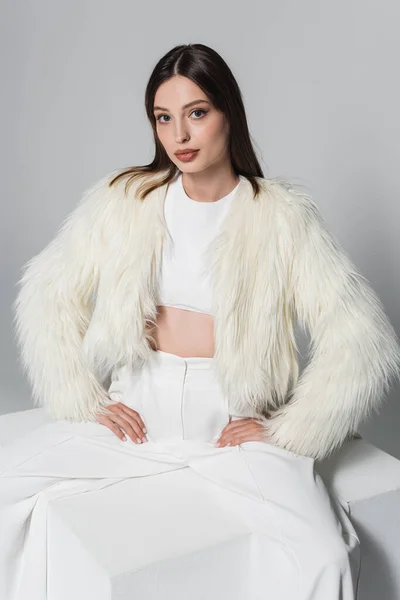 Jovem mulher na moda casaco de pele falso e roupa branca total sentado com as mãos sobre os quadris no cubo isolado em cinza — Fotografia de Stock