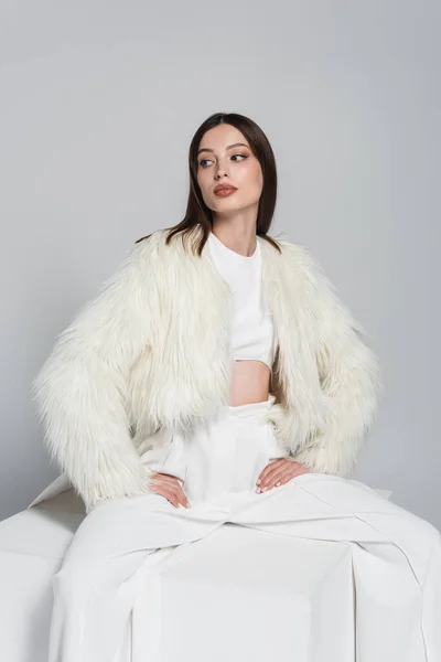 Mulher elegante em casaco de pele de falso branco sentado com as mãos sobre os quadris no cubo isolado no cinza — Stock Photo