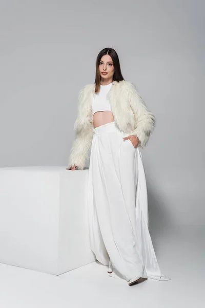 Full length of trendy woman in trendy faux fur jacket and total white outfit standing with hand in pocket near cube on grey — Stock Photo