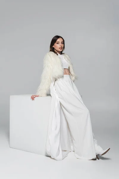 Полная длина молодой женщины в белом искусственном меховом пиджаке и модном наряде, стоящем рядом с кубом на сером — стоковое фото