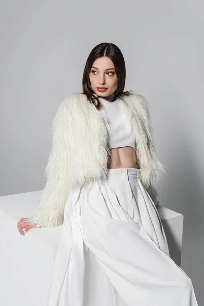 Модная молодая женщина в искусственном меховом пиджаке и полном белом наряде, опирающаяся на куб на сером — стоковое фото