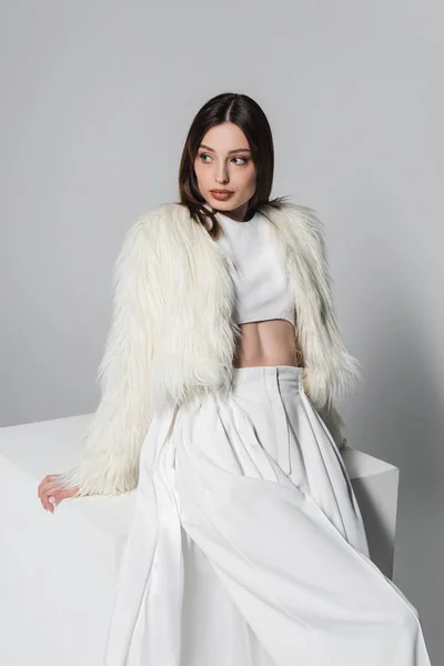 Mujer joven de moda en chaqueta de piel sintética y traje blanco total apoyado en el cubo en gris - foto de stock