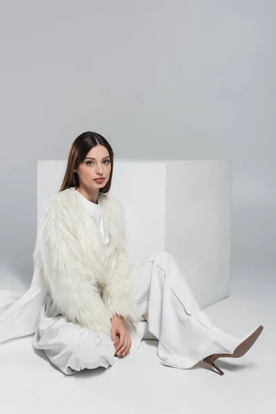 Полная длина модной женщины в искусственной меховой куртке и полный белый наряд сидит рядом с кубом на сером — стоковое фото