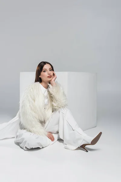 Full length of young woman in faux fur jacket and total white outfit sitting near cube on grey — Stock Photo