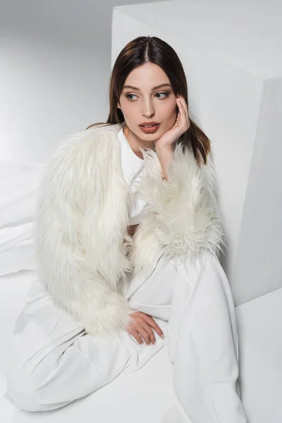 Visão de alto ângulo da jovem mulher em casaco de pele falso e roupa branca total sentada perto do cubo no cinza — Fotografia de Stock
