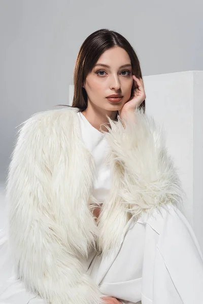 Young woman in white faux fur jacket looking at camera near cube isolated on grey — Stock Photo