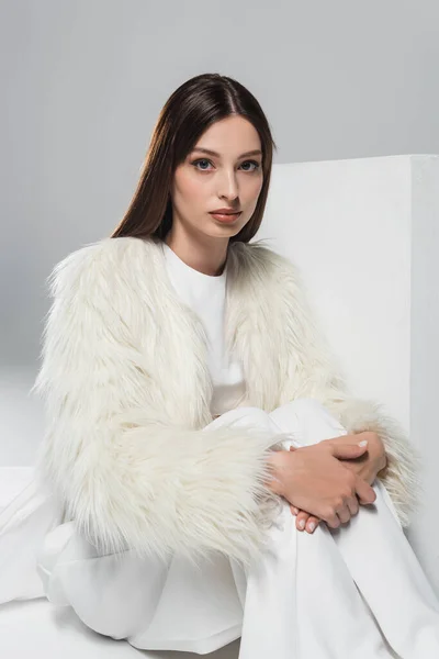 Stylish young woman in white faux fur jacket looking at camera near cube on grey — Stock Photo