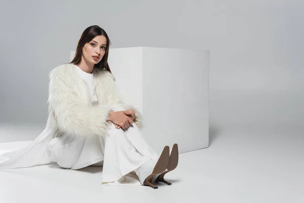 Полная длина стильной молодой женщины в белом искусственном меховом пиджаке, смотрящей на камеру рядом с кубом на сером фоне — стоковое фото