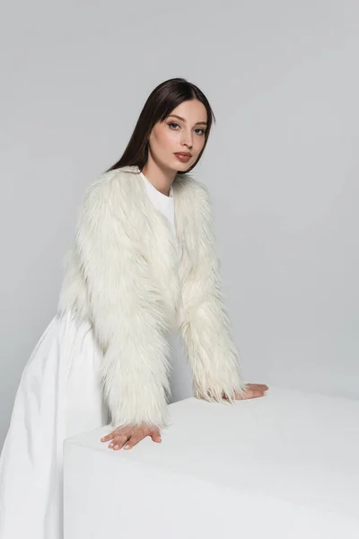 Young woman in faux fur jacket looking at camera and leaning on white cube isolated on grey — Stock Photo