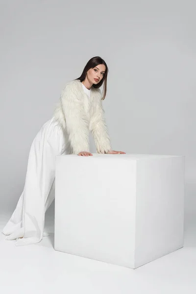 Полная длина стильной молодой женщины в искусственном меховом пиджаке, смотрящей в камеру, опираясь на белый куб на сером — стоковое фото