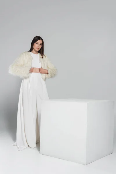 Piena lunghezza di donna elegante in giacca di pelliccia sintetica guardando la fotocamera mentre in piedi vicino cubo bianco sul grigio — Foto stock
