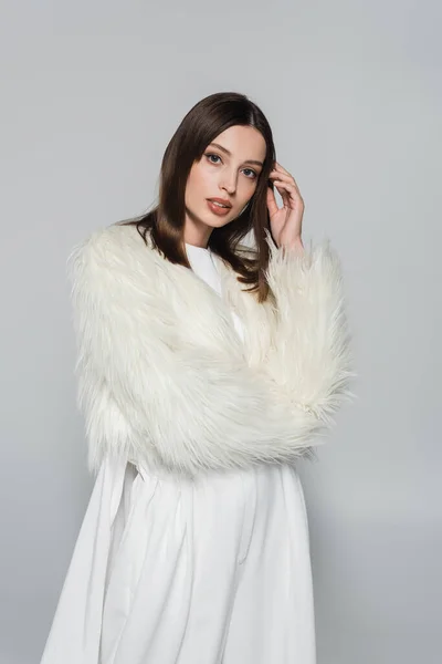 Retrato de mulher jovem elegante em casaco de pele de falso branco olhando para a câmera isolada no cinza — Fotografia de Stock
