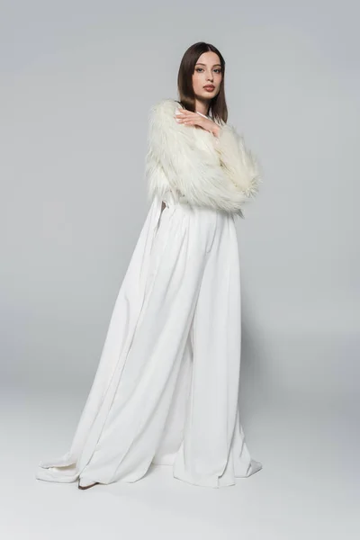 Full length of trendy woman in totally white outfit and faux fur jacket posing on grey — Stock Photo