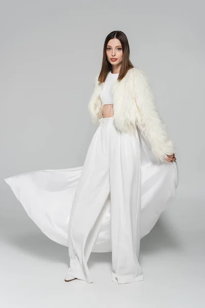 Full length of trendy woman in totally white outfit and faux fur jacket standing on grey — Stock Photo
