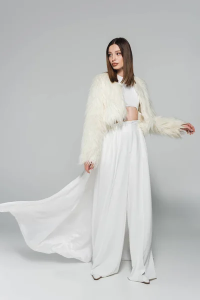 Full length of young woman in totally white outfit and trendy faux fur jacket posing on grey — Stock Photo