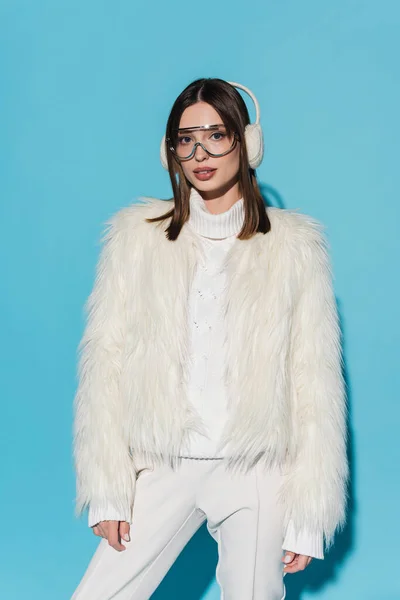 Elegante giovane donna in paraorecchie e occhiali in posa in giacca di pelliccia ecologica su blu — Foto stock