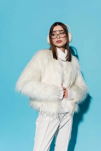 Trendy woman in earmuffs and eyeglasses posing in faux fur jacket while looking away on blue — Stock Photo