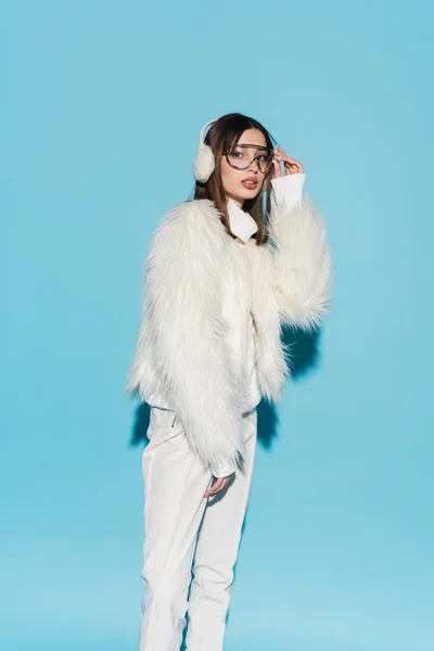 Stylish woman in earmuffs adjusting eyeglasses while posing in faux fur jacket on blue — Stock Photo