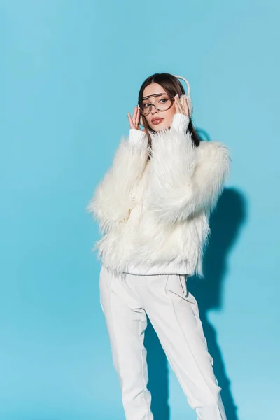 Stylish young woman in earmuffs adjusting trendy eyeglasses while posing in faux fur jacket on blue — Stock Photo