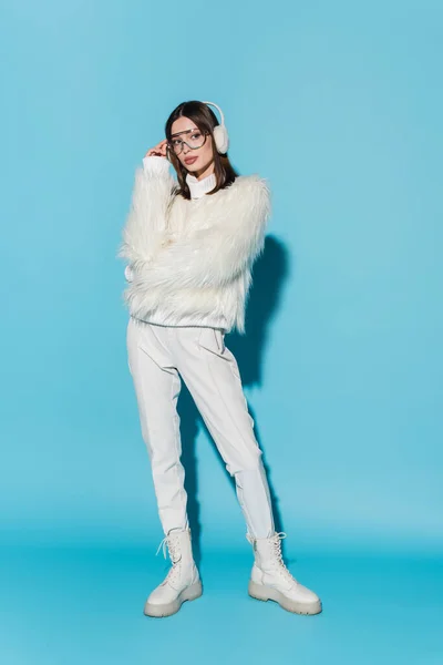 Full length of stylish young woman in earmuffs adjusting trendy eyeglasses while posing in faux fur jacket on blue — Stock Photo