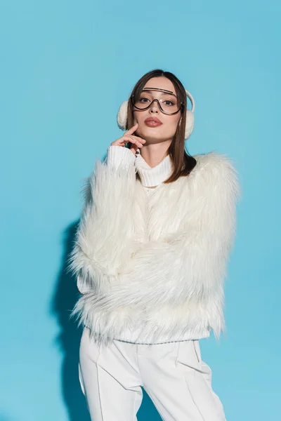 Stylish young woman in earmuffs and trendy eyeglasses posing in faux fur jacket on blue — Stock Photo