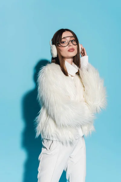 Stylish young woman adjusting winter earmuffs while posing in white faux fur jacket on blue — Stock Photo
