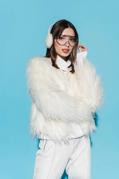 Stylische junge Frau mit Winterohrenschützer und Brille posiert in weißer Kunstpelzjacke auf blauem Grund — Stockfoto