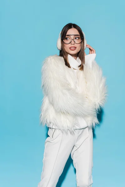 Stylish young woman in winter earmuffs and trendy eyeglasses posing in white faux fur jacket on blue — Stock Photo