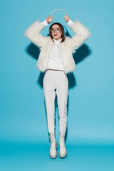 Full length of stylish woman in eyeglasses holding earmuffs and levitating on blue — Stock Photo