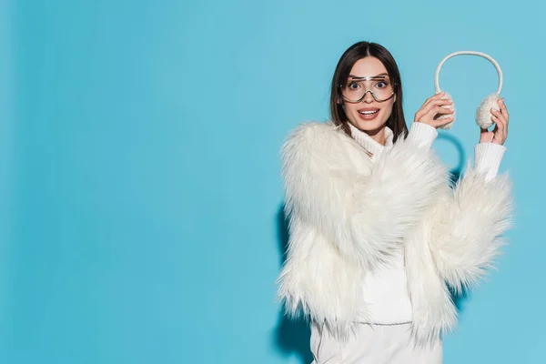 Amazed young woman in eyeglasses and stylish faux fur jacket holding winter earmuffs on blue — Stock Photo