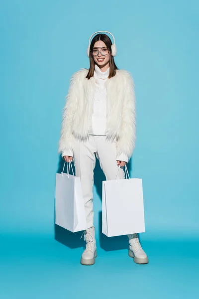 Full length of happy woman in earmuffs and eyeglasses holding shopping bags on blue — Stock Photo