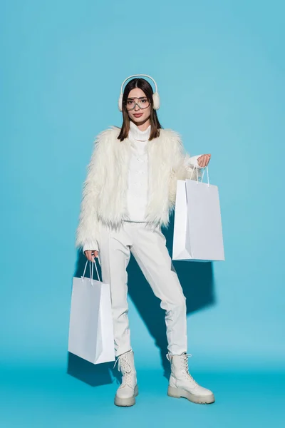 Full length of stylish woman in earmuffs and eyeglasses holding shopping bags on blue — Stock Photo