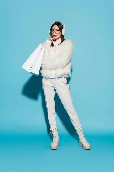 Full length of stylish woman in faux fur jacket and eyeglasses holding shopping bags on blue — Stock Photo