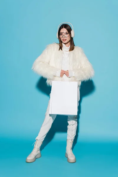 Piena lunghezza di donna elegante in paraorecchie e giacca di pelliccia sintetica che tiene borse della spesa su blu — Foto stock