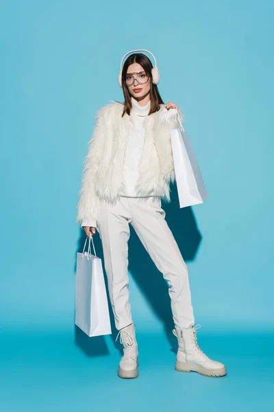 Longitud completa de la mujer joven en orejeras y chaqueta de piel sintética elegante celebración de bolsas de compras en azul - foto de stock