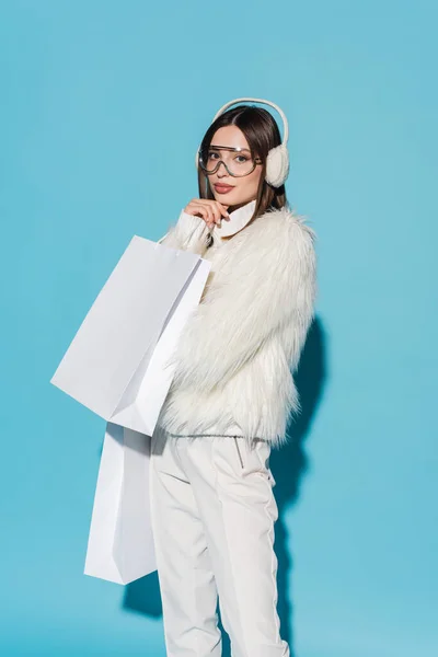 Young woman in winter earmuffs and trendy faux fur jacket holding shopping bags on blue — Stock Photo