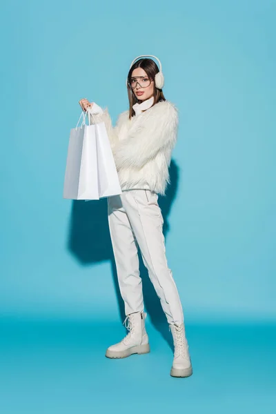 Longitud completa de la mujer bonita en orejeras de invierno y chaqueta de piel sintética elegante celebración de bolsas de compras en azul - foto de stock