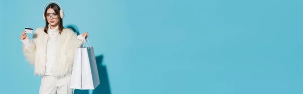 Jeune femme en cache-oreilles d'hiver et veste élégante en fausse fourrure tenant carte de crédit et sacs à provisions sur bleu, bannière — Photo de stock