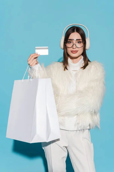 Young woman in eyeglasses and stylish faux fur jacket holding credit card and shopping bags on blue — Stock Photo