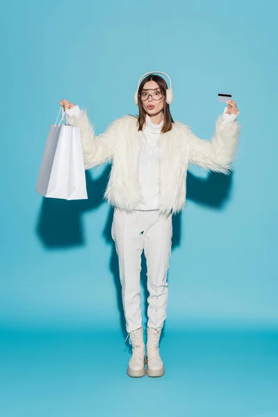Comprimento total da mulher surpreendida em óculos e casaco de pele falso elegante segurando cartão de crédito e sacos de compras em azul — Fotografia de Stock