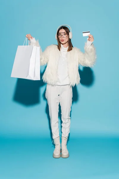 Full length of surprised woman in earmuffs and white faux fur jacket holding credit card and shopping bags on blue — Stock Photo
