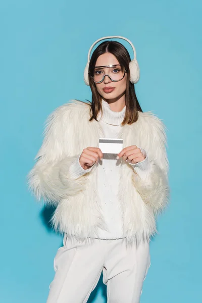 Jeune femme en lunettes et veste élégante en fausse fourrure tenant carte de crédit sur bleu — Photo de stock