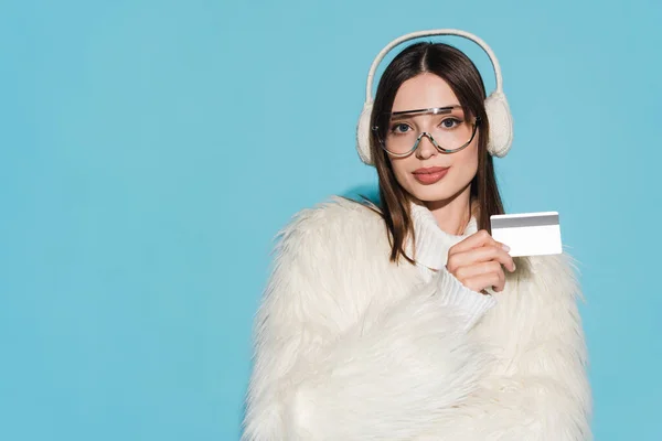 Pleased young woman in eyeglasses and stylish faux fur jacket holding credit card isolated on blue — Stock Photo