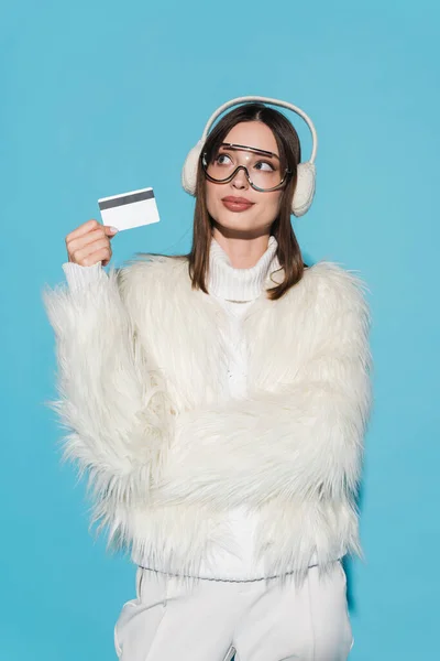 Pensive young woman in eyeglasses and stylish faux fur jacket holding credit card isolated on blue — Stock Photo