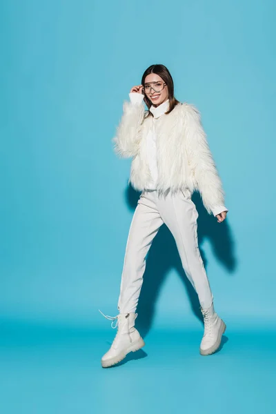 Full length of cheerful woman in faux fur jacket and stylish eyeglasses walking on blue — Stock Photo