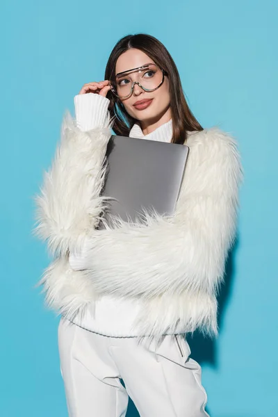 Jeune femme dans des lunettes de vue à la mode et veste élégante en fausse fourrure tenant ordinateur portable sur bleu — Photo de stock