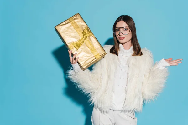Stylische Frau in Kunstpelzjacke und Brille mit Weihnachtsgeschenk auf blauem Hintergrund — Stockfoto