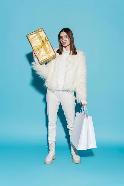 Pleine longueur de femme élégante en fausse veste de fourrure et lunettes tenant cadeau de Noël et sacs à provisions sur bleu — Photo de stock