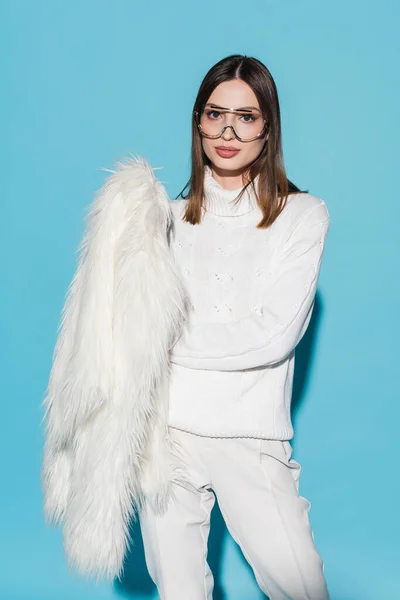 Stylish young woman in trendy eyeglasses holding white faux fur jacket on blue — Stock Photo